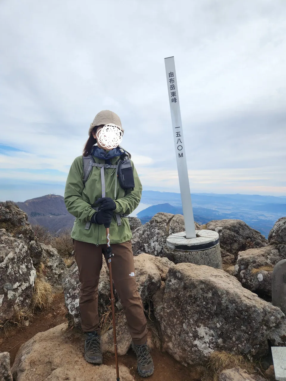 12月の由布岳東峰の山頂で、アルパインライトパンツをはいて標柱の横に立つ